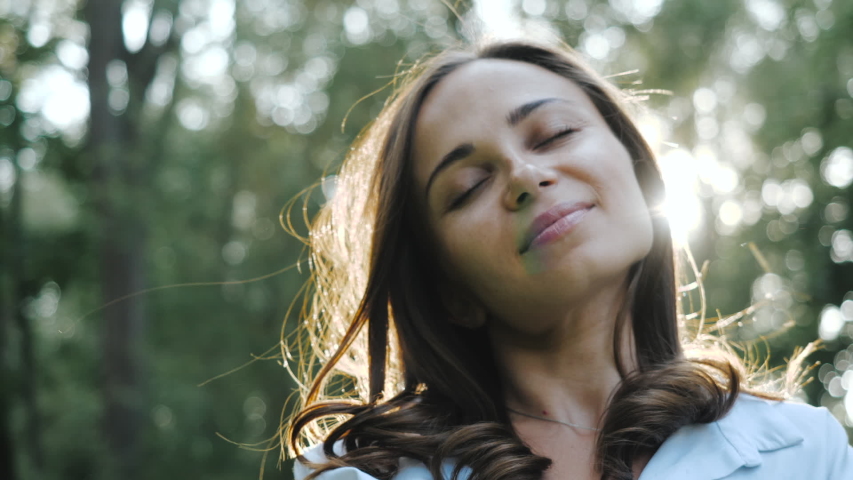 Close-up view of the face of a pretty smiling girl in a park among the trees against the backdrop of a bright sun. A spring breeze blows lightly at the girl. Woman closed her eyes and enjoys the air.