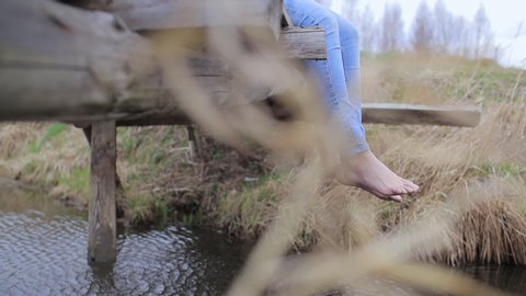 Woman legs in jeans without shoe dangling from old wooden bridge, close-up view