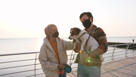 Young happy couple in protective medical masks playing with cute Jack Russel terrier dog outdoors near the sea during sunrise