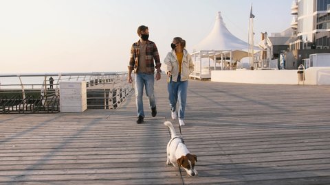 Young happy couple in protective medical masks walking with cute Jack Russel terrier dog outdoors near the sea, slow motion