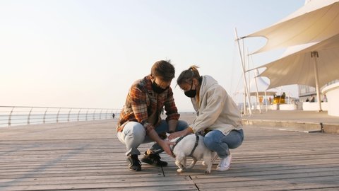 Young happy couple in protective medical masks petting cute Jack Russel terrier dog outdoors near the sea, slow motion