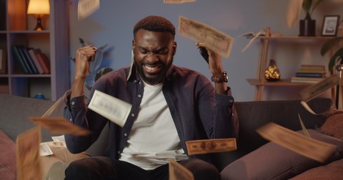 Handsome young man using smartphone and winning big money on sports betting. Excited guy sitting on sofa and rejoicing victoryb while money falling from above. Home background.