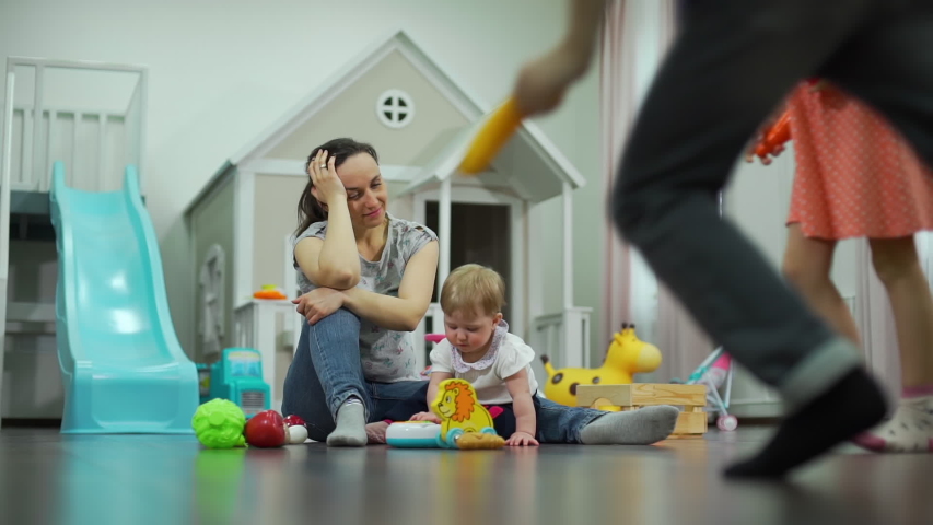 Naughty Children Running Around Distracted Mother in Messy Nursery. Tired Young Woman Sitting on the Floor with Infant Child. Concept of Family and Lifestyle Royalty-Free Stock Footage #1051567705