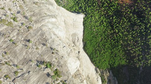 Downgoing view from the edge of the top of famous El Capitan rock on its vertical granite walls steeply descending to the ground, which is covered with dense forest. Yosemite National Park. Aerial, 4K