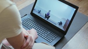 A man sits on a yoga mat at home on self-isolation, watches yoga classes online using a laptop, a girl trainer shows asanas.