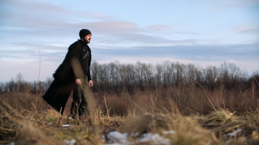 Bearded man in black leather trousers and fur coat mantle walking like warrior in winter steppe, Ukraine. Grass is dry and light is sunny and warm, sky is blue