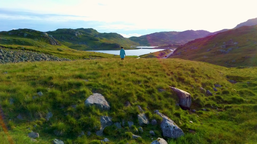 Aerial drone flight past a young girl standing on a hill in the middle of a beautiful valley. Road passes stones and green hills in area of lake Ivlungsvotni in Norway. Taken in the summer at sunset