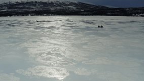 Aerial video on flying forward above wet ice lake with unidentified people driving snowmobiles and fishing. Melting water at ice reflecting Sun and dark blue sky
