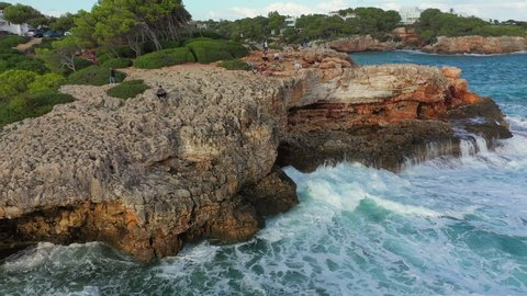 Aerial panning shot of people walking on rocky cliff in sea against sky, drone flying around tourists by waves splashing near city - Cales de Mallorca, Spain