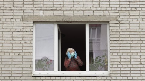 Old woman in panics stay at window in protection mask. Checks her temperature. Isolated at home on quarantine. Virus outbreak. Elder grandmother. COVID-19 pandemic coronavirus. Social distancing