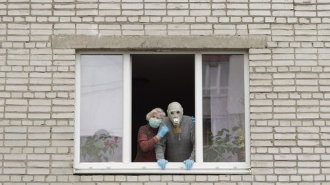Old grandfather in gas mask and grandmother in protection mask stay at window. Isolated at home on quarantine. Virus outbreak. Sad elder man, woman. COVID-19 pandemic coronavirus. Social distancing