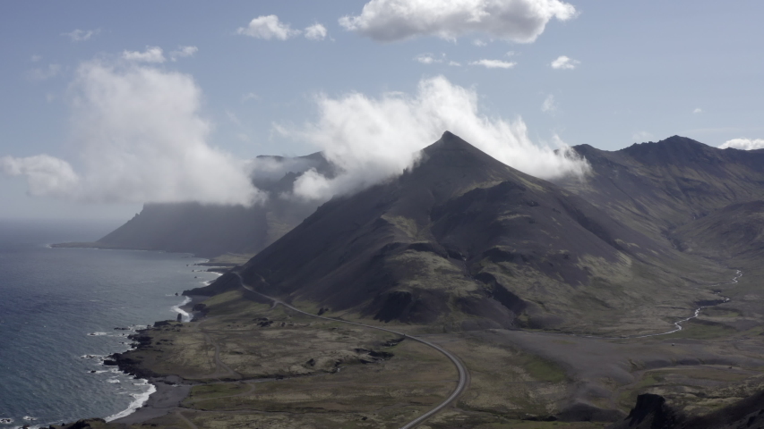 Aerial shot of clouds covering mountain peak by sea, drone flying forward over coastal landscape - Southwestern Iceland, Iceland