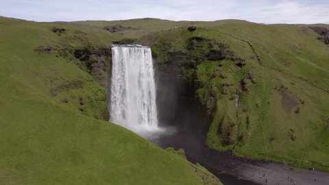 Aerial panning shot of beautiful waterfall by tourists on green cliff, drone descending over river amidst moss covered rock formations - Southwestern Iceland, Iceland