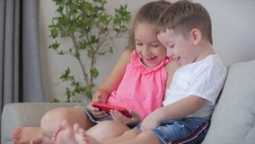 Happy children playing at home relaxing use a smartphone cuddling sit on sofa daughter and son,look at the screen of a cell phone, watch cartoons.