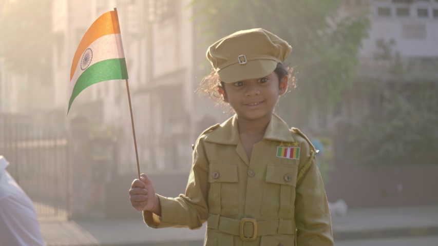 A young smiling, happy cute, adorable kid, girl child wearing military, army, soldier costume, dress,  uniform and holding an Indian national, tricolour flag in hand standing outside in Mumbai, India 