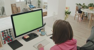 Young woman at home works on computer with green mock-up screen. Girl in cozy clothes sitting in living room. Over shoulder camera shot. 4k video footage slow motion