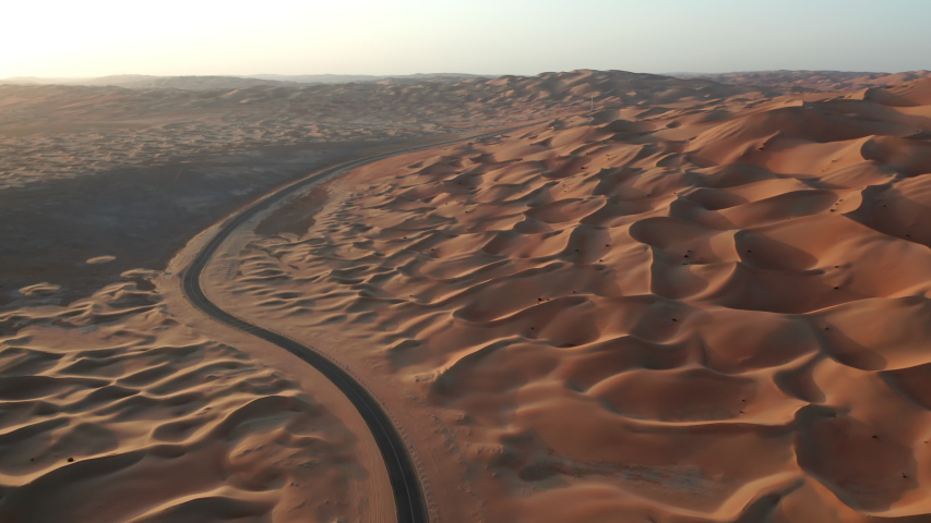 Aerial shot of an empty road in Abu-Dhabi Liwa desert region. Sunrise time with warm vivid colors. Drone flying forward with camera tilts up from road to sand dunes Royalty-Free Stock Footage #1051608304