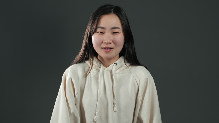 Smiling asian woman with long hair holding finger on her lips over grey background. Gesture of shhh, secret, silence. Close up.