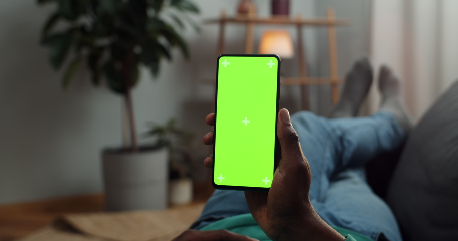 Young man lying on couch with modern smartphone in his hand while looking on mock up screen with trecking markers on it. Concept of green screen and chroma key. Cosy home background Royalty-Free Stock Footage #1051618927