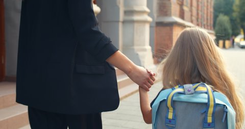 Close up view of little blond girl in uniform with bag walking with her mother hand in hand. Backside view of child holding her mom and entering in school early morning. Outdoors
