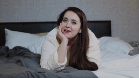 A woman lying in bed speaks to the camera by video link while having video chat on a smartphone. Stay at home, videoconference, modern technology. Model plus size