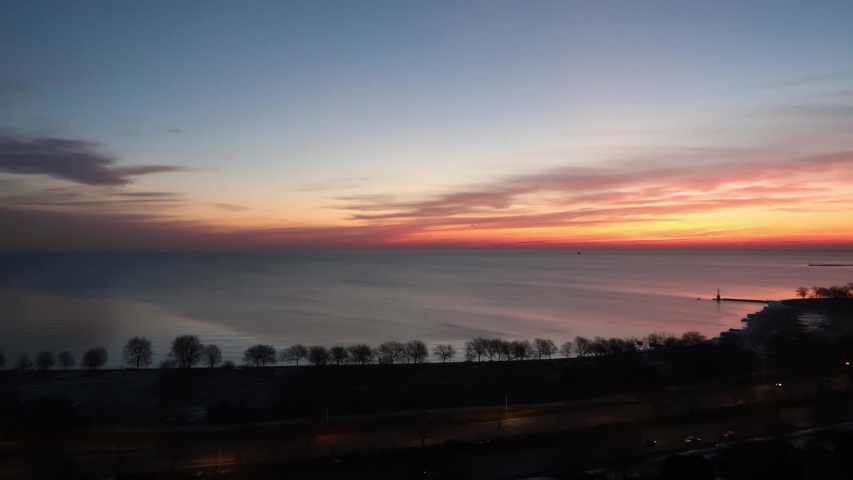 Aerial timelapse of a beautiful soft and feathery pink, orange, blue and yellow sky sunrise reflecting on the water of Lake Michigan in Chicago with tree lined shoreline below.