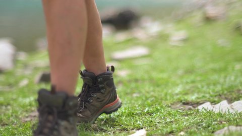 Hiker girl foots in nature with boots. Outdoor woman strolling by the mountain lake in natural terrain. Trekking and hiking in lakeside. Step pace footstep meadow grass pasture lawn. Grassland nature.