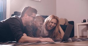 Happy family young parents couple and cute kid son using laptop looking at computer screen enjoying watching funny social media video doing online shopping relaxing on carpet at home together