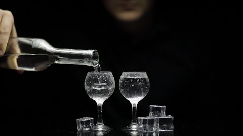 Bartender pouring up frozen vodka from a bottle into two shots glasses with ice cubes against black background. Barman pour of clear transparent alcohol drink rum tequila in shot-glass. Slow motion
