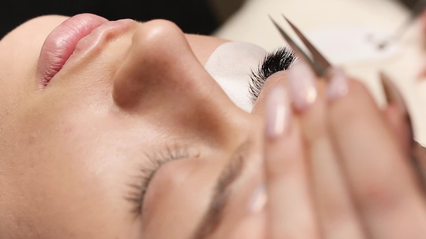 Eyelashes extensions. Fake eyelashes. Eyelash extension procedure.Close up portrait of woman eye with long eyelashes. Professional stylist lengthening female lashes. Master and client in beauty salon. | Shutterstock HD Video #1051650136