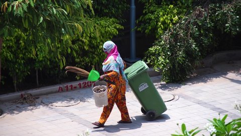 Gurgaon, Delhi, India - circa 2020 : Old lady in saree and mask cleaning pavements sidewalks and streets with a wooden broom and putting garbage into a green movable dustbin. Shows the agressive