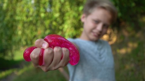 Closeup view video portrait of young happy healthy white kid holding pink or red sparkling transparent slime in hands, playing it happily, squeezing and pulling it. 