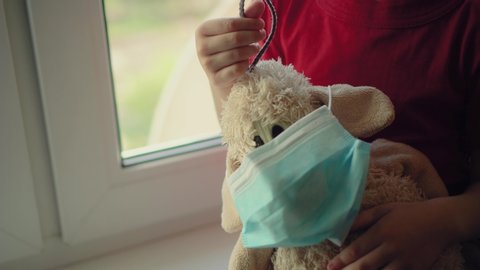 Sad illness child on home quarantine. Boy and his teddy bear both in protective medical masks sits on windowsill and looks out window. Virus protection, coronavirus pandemic, prevention epidemic.