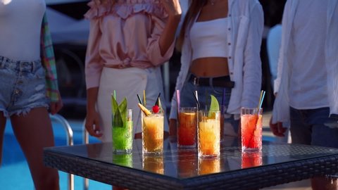 Group of friends having fun at poolside summer party taking colorful cocktails from table and clinking glasses smiling. People toast drinking fresh juice at luxury tropical villaon summer day.