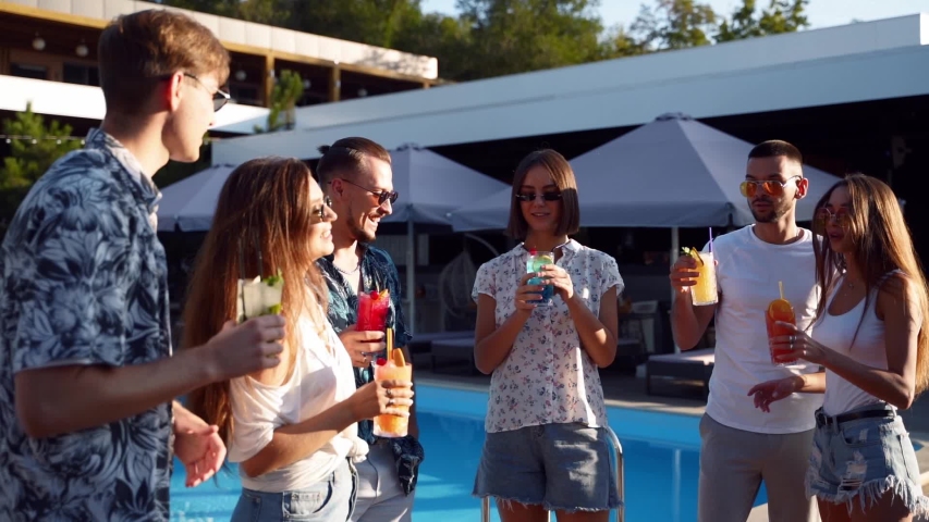 Group of friends having fun at poolside summer party clinking glasses with summer cocktails on sunny day near swimming pool. People toast drinking fresh juice at luxury villa on tropical vacation. | Shutterstock HD Video #1051669390