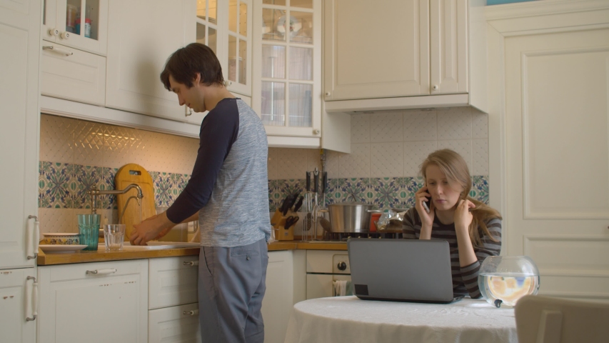 Portrait of a young woman working from home at the kitchen. Her husband washing dishes. Daily family life, remote job.