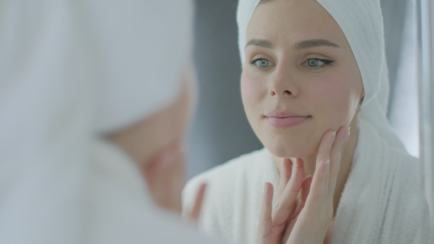 Close up pretty brunette woman in a towel on her head touching face and looking at mirror. Skin care and beauty health concept
 | Shutterstock HD Video #1051686010