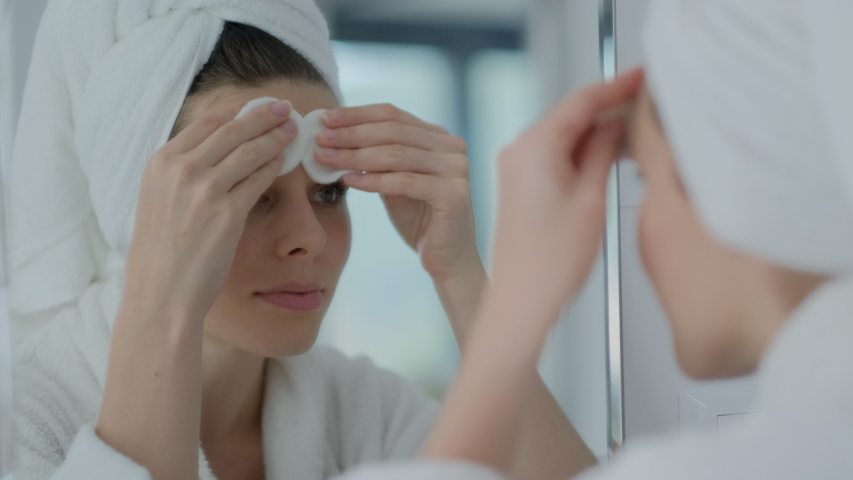 Attractive Young brunette woman in a towel on her head cleaning the face with a cotton pad looking at mirror, removing make up.
 | Shutterstock HD Video #1051686193