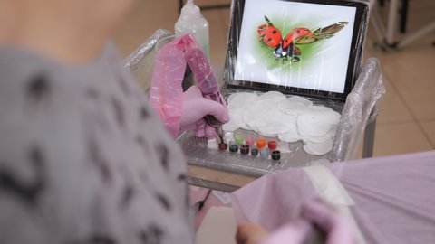 A tattoo master dips a tattoo machine with needles in red ink. The tattooist is preparing to put a tattoo on the girl's skin, in the background is a tablet with a picture of a ladybug.