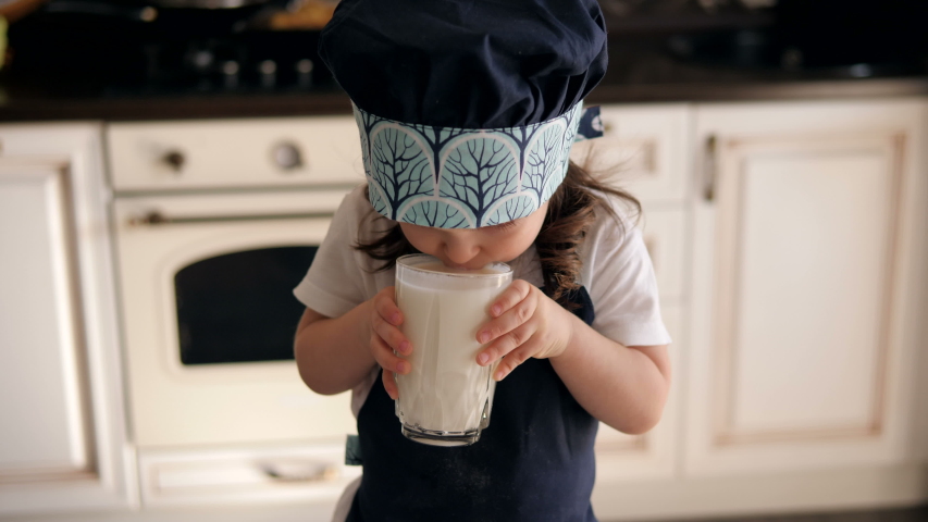 Portrait of a little three-year-old girl in an apron and cap in the kitchen, she drinks kefir or milk and leaves a milky moustache on her lips. There are Christmas cookies on the table. Royalty-Free Stock Footage #1051687168