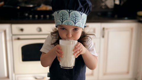 Portrait of a little three-year-old girl in an apron and cap in the kitchen, she drinks kefir or milk and leaves a milky moustache on her lips. There are Christmas cookies on the table.