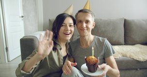 Happy couple of bloggers in birthday caps recording video and blowing out a candle on a chocolate cake. Married couple celebrating anniversary and talking with friends on video during self-isolation