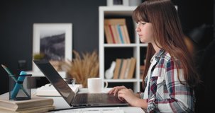 Side view of teenager girl in checkered shirt using laptop for remote studying at home. Cute little female sitting at table and doing homework on computer.