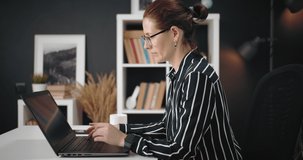 Side view of concentrated mature woman with hair knot sitting at table and using personal laptop for remote work. Concept of business career and isolation