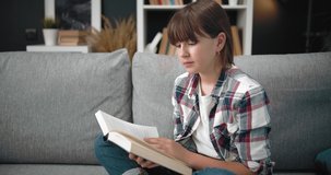 Pretty young girl in casual clothing sitting on grey couch and reading interesting book. Female teenager with dark hair spending free time at home for education.