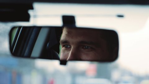 Young man inside car. Happy positive caucasian man driving car fast and look at rear view mirror with smile. Blurred background. Slow motion.