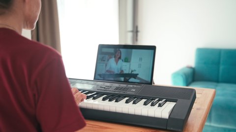A young student watching music lesson online and studying from home. Young woman practicing her piano skills while looking at computer screen following professor on a video call. 
