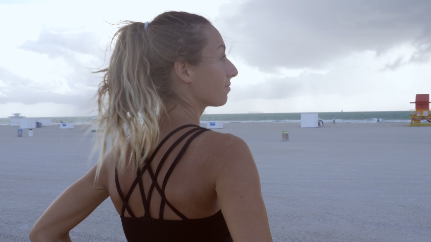 Sportive young woman on beach contemplating ocean and tidying pony tail, Miami Beach