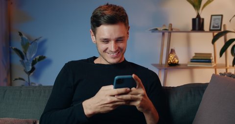 Cheerful millennial man communicating with friends in social networks and laughing while getting funny message. Handsome young guy looking at screen and typing while sitting on sofa