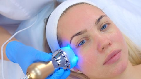 Portrait of woman on apparatus hardware phonophoresis face procedure with hyaluronic acid gel in cosmetology clinic, portrait closeup. Cosmetologist doctor moving manipula with cooling blue light.
